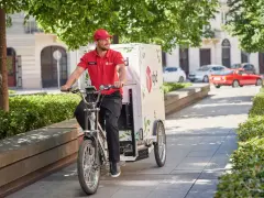 Uśmiechnięty kurier rowerowy DPD w czerwonej koszulce jedzie ulicą z zielonymi drzewami.