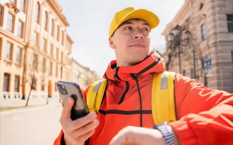 Kurier Glovo w żółtej czapce i pomarańczowej kurtce sprawdza telefon, gotowy do odbioru płatności Glovo cash.