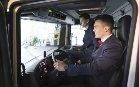Dwóch młodych mężczyzn w garniturach ćwiczy jazdę ciężarówką na symulatorze. Zastanawiają się, ile zarabia kierowca na godzinę.