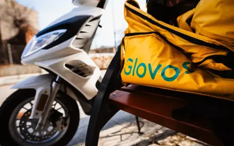 Żółta torba z logo Glovo na ławce obok białego skutera. Czeka na kolejnego kuriera Glovo.