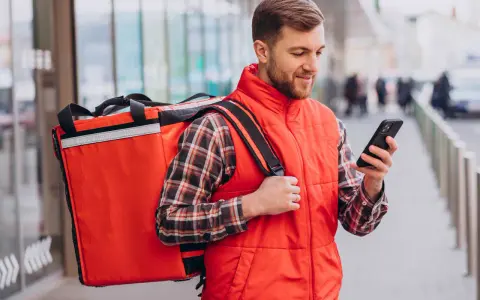 Uśmiechnięty dostawca jedzenia z dużą czerwoną torbą na ramieniu sprawdza trasę na telefonie.