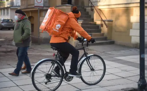 Kurier rowerowy w pomarańczowej kurtce z plecakiem Pyszne.pl, zarobki zależą od liczby dostaw.
