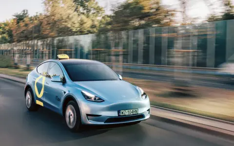 Niebieski Tesla Model Y z żółtym znakiem taxi na dachu i logo iTaxi na drzwiach, spełniający wymagania samochodu dla tej usługi.