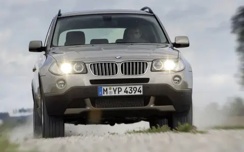 Złoty SUV BMW X3 z włączonymi światłami jedzie po szutrowej drodze, unosząc kurz. To jeden z najbardziej komfortowych aut na drodze.