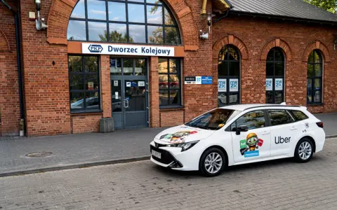 Biały samochód Ubera zaparkowany przed dworcem kolejowym. Kierowca z własnym samochodem czeka na pasażerów.