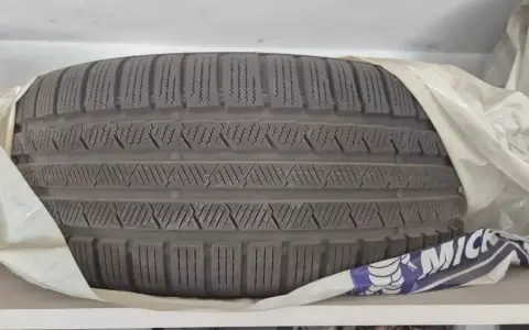 Opona leży poziomo, owinięta w biały materiał. Widać bieżnik z charakterystycznymi nacięciami.