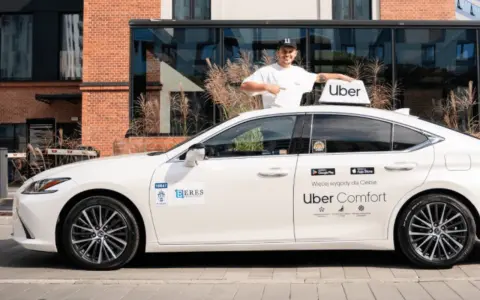 Mężczyzna w czapce z daszkiem stoi obok białego samochodu z logo Uber. To część floty Uber, gotowa do jazdy.