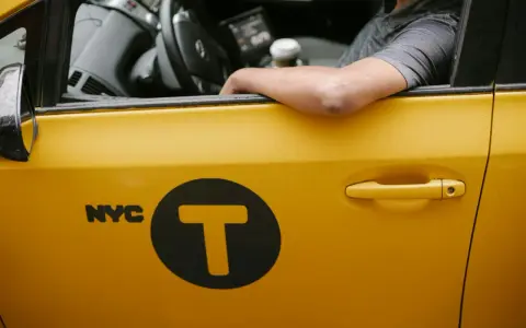 Kierowca żółtej taksówki NYC z logo "T" w kole. Zastanawiasz się, ile zarabia taksówkarz w tym mieście?