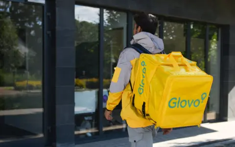 Kurier Glovo pieszo idzie z żółtą torbą na plecach.