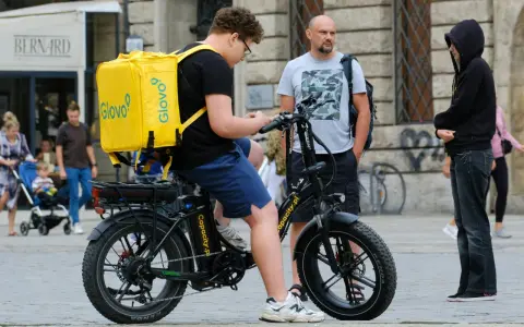 Młody kurier Glovo na rowerze elektrycznym sprawdza telefon. Obok stoi mężczyzna, a w tle widać ludzi i budynki.