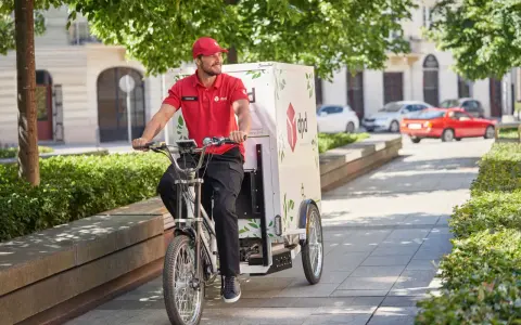 Uśmiechnięty kurier rowerowy DPD w czerwonej koszulce jedzie ulicą z zielonymi drzewami.