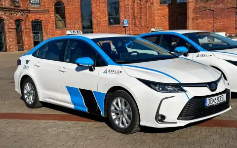 Białe taksówki z niebieskimi pasami i logo Avalon, spełniające uber wymagania samochodu, stoją przed ceglanym budynkiem.