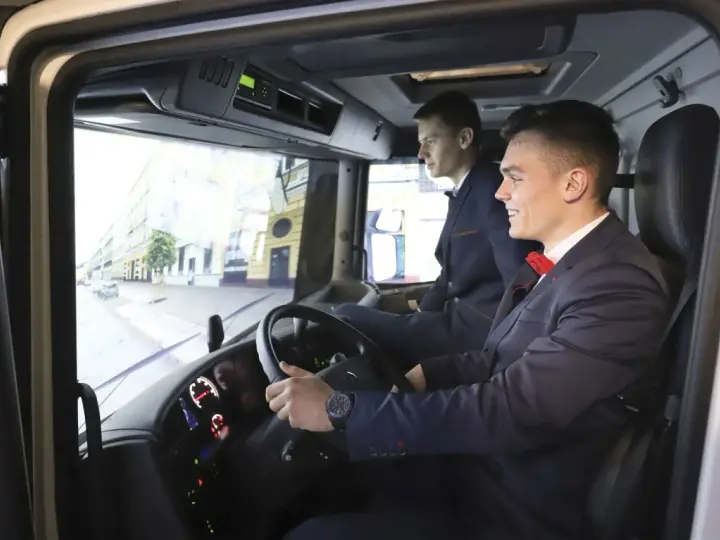 Dwóch młodych mężczyzn w garniturach ćwiczy jazdę ciężarówką na symulatorze. Zastanawiają się, ile zarabia kierowca na godzinę.