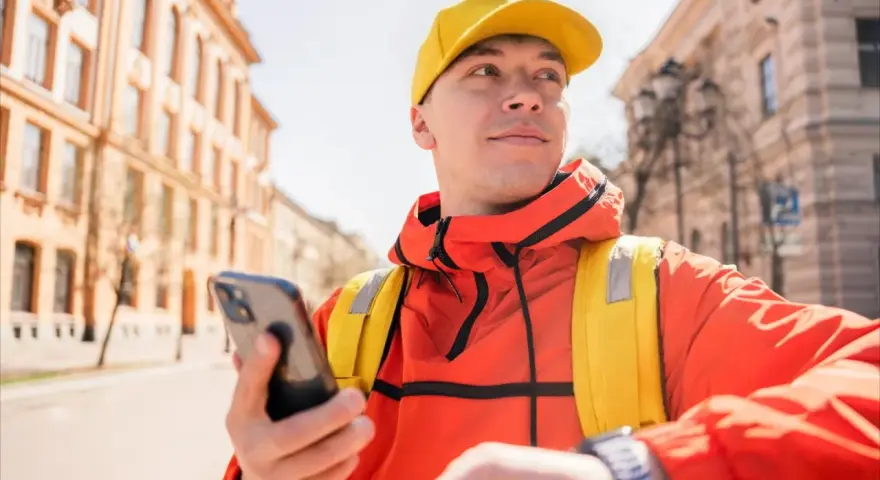 Kurier Glovo w żółtej czapce i pomarańczowej kurtce sprawdza telefon, gotowy do odbioru płatności Glovo cash.
