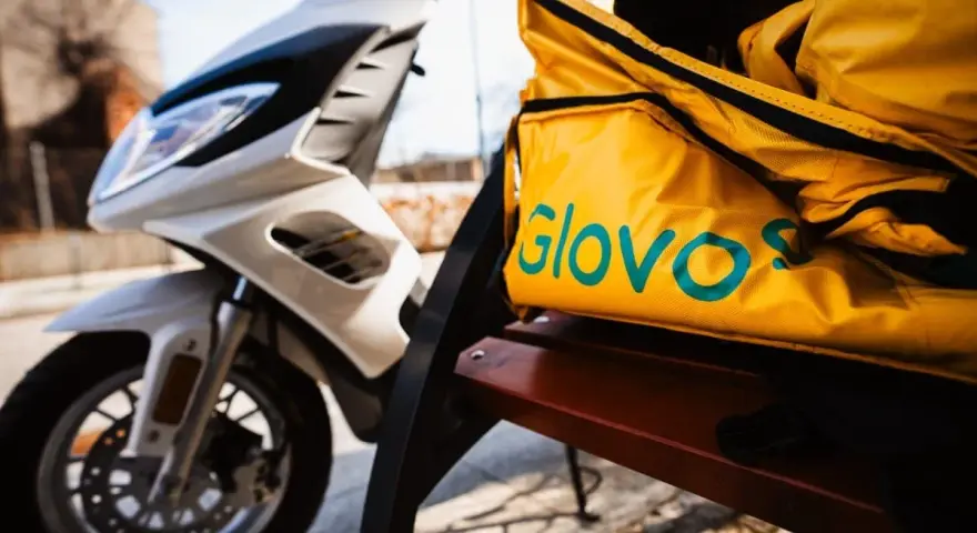 Żółta torba z logo Glovo na ławce obok białego skutera. Czeka na kolejnego kuriera Glovo.