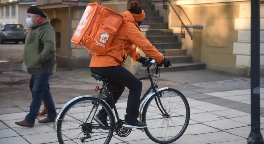 Kurier rowerowy w pomarańczowej kurtce z plecakiem Pyszne.pl, zarobki zależą od liczby dostaw.