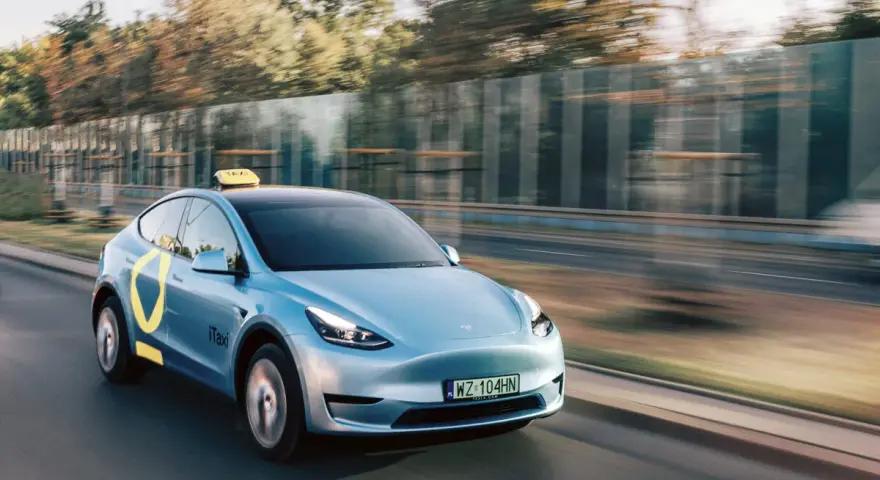 Niebieski Tesla Model Y z żółtym znakiem taxi na dachu i logo iTaxi na drzwiach, spełniający wymagania samochodu dla tej usługi.