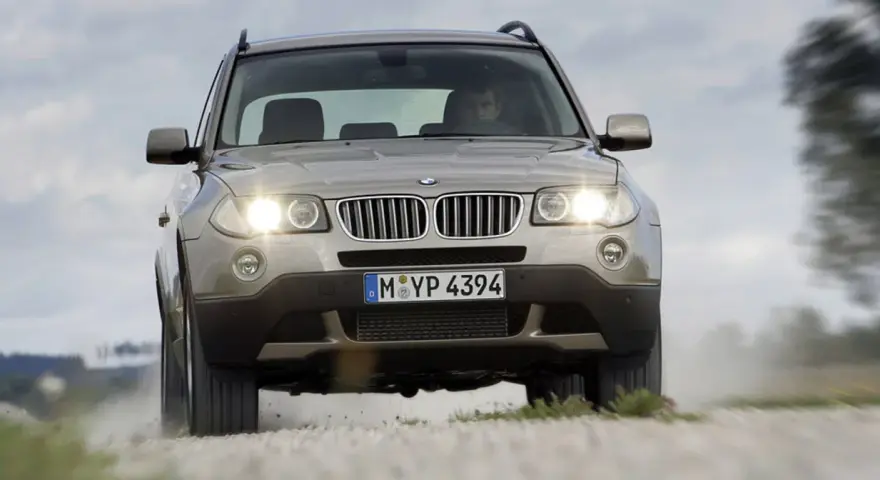 Złoty SUV BMW X3 z włączonymi światłami jedzie po szutrowej drodze, unosząc kurz. To jeden z najbardziej komfortowych aut na drodze.