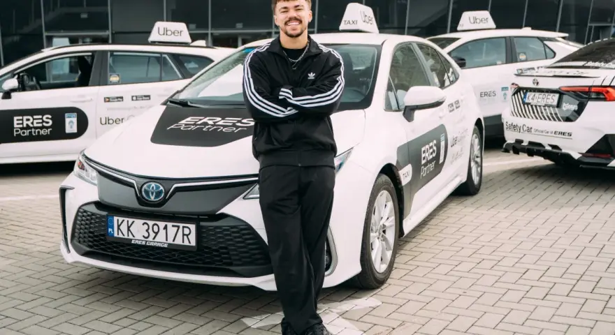 Młody mężczyzna w dresie stoi przed białymi samochodami Uber z napisem "ERES Partner". To jego praca prywatny kierowca.