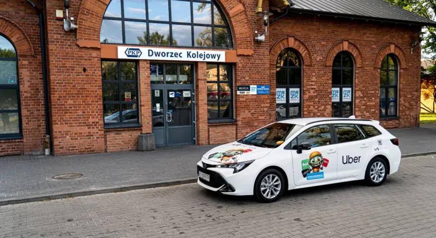 Biały samochód Ubera zaparkowany przed dworcem kolejowym. Kierowca z własnym samochodem czeka na pasażerów.