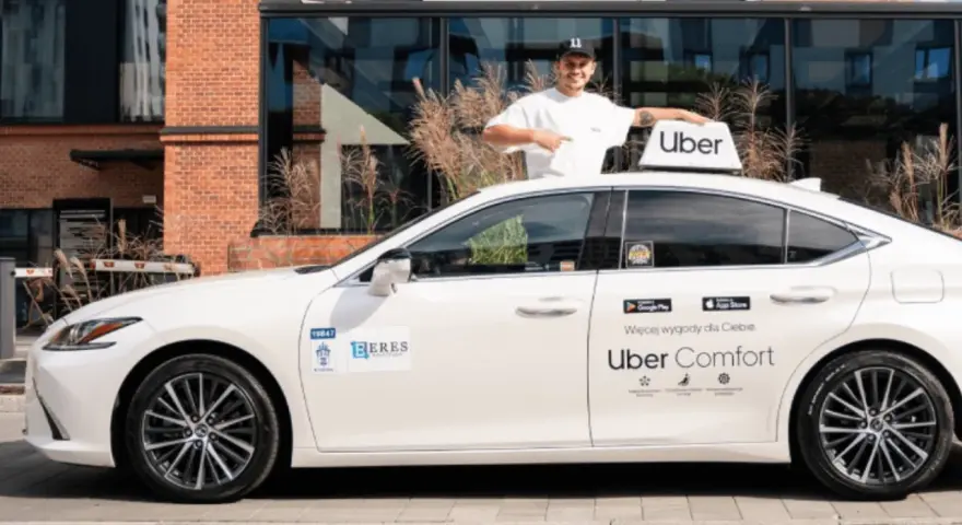 Mężczyzna w czapce z daszkiem stoi obok białego samochodu z logo Uber. To część floty Uber, gotowa do jazdy.
