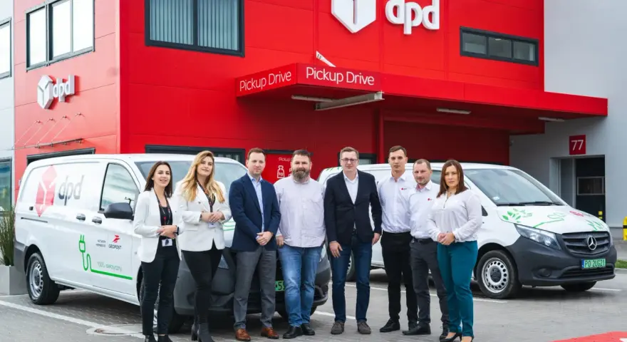 Zespół DPD pozuje przed budynkiem z logo firmy i elektrycznym dpd bus.