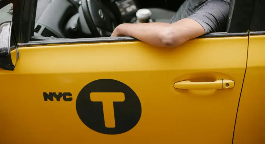 Kierowca żółtej taksówki NYC z logo "T" w kole. Zastanawiasz się, ile zarabia taksówkarz w tym mieście?
