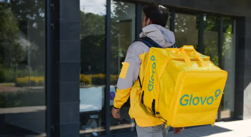 Kurier Glovo pieszo idzie z żółtą torbą na plecach.