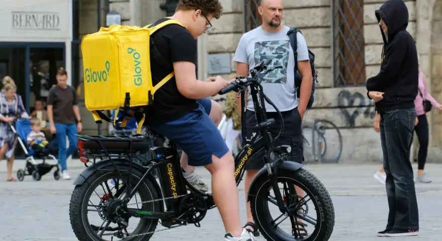 Młody kurier Glovo na rowerze elektrycznym sprawdza telefon. Obok stoi mężczyzna, a w tle widać ludzi i budynki.