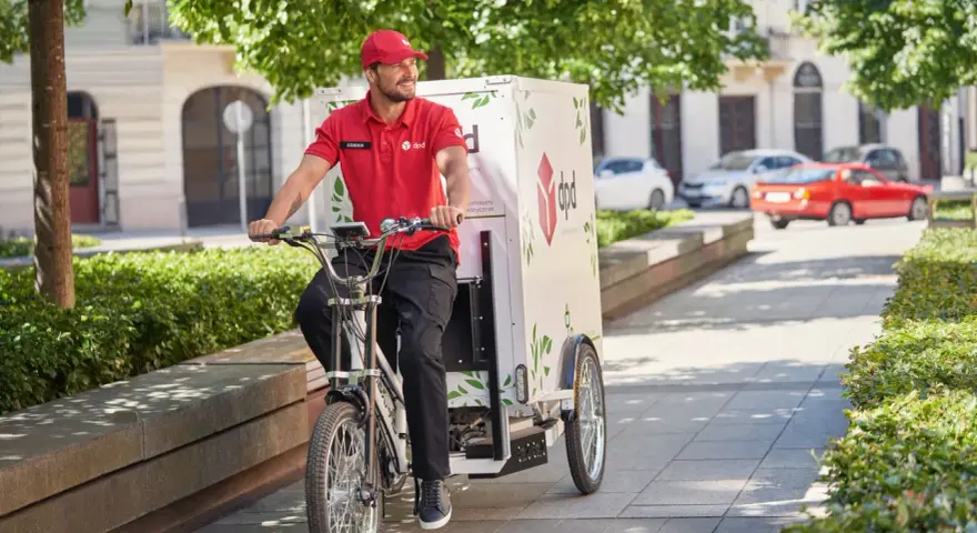 Uśmiechnięty kurier rowerowy DPD w czerwonej koszulce jedzie ulicą z zielonymi drzewami.