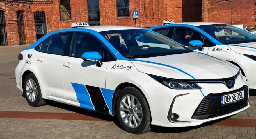 Białe taksówki z niebieskimi pasami i logo Avalon, spełniające uber wymagania samochodu, stoją przed ceglanym budynkiem.