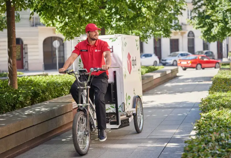 Uśmiechnięty kurier rowerowy DPD w czerwonej koszulce jedzie ulicą z zielonymi drzewami.